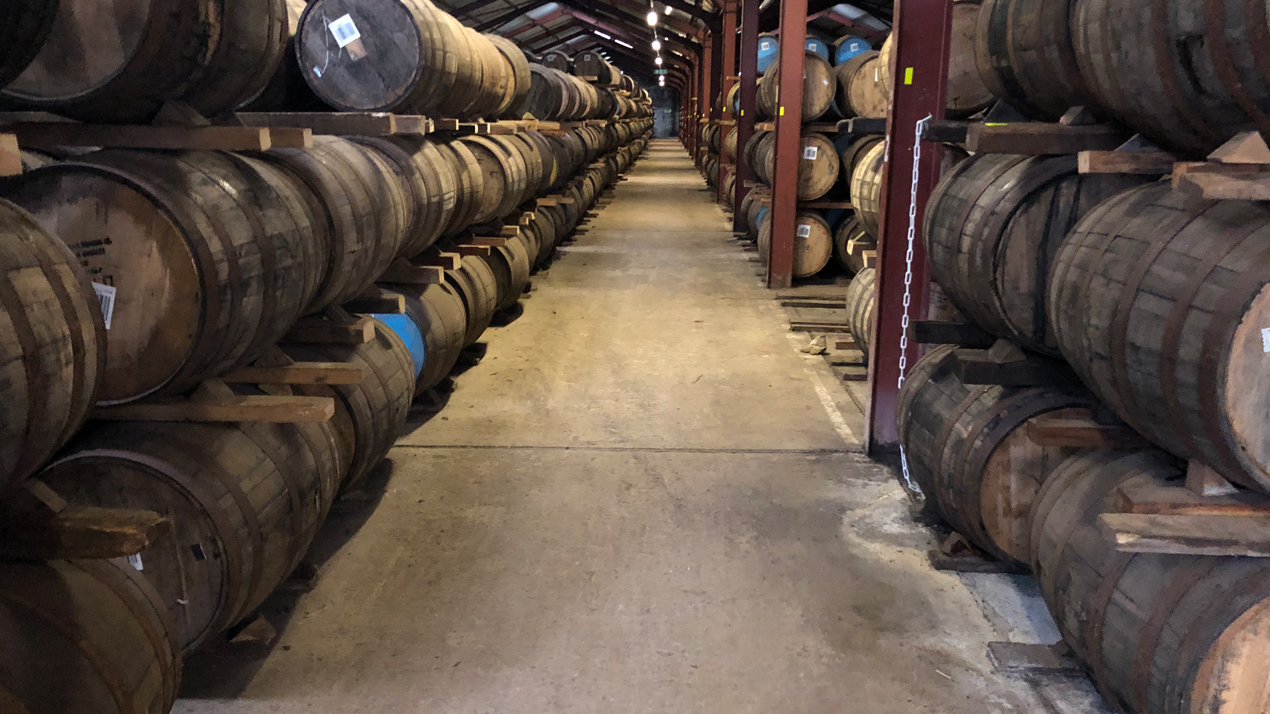 Långa rader av ekfat med whisky, tre rader högt, sträcker sig bortåt i mörk lagerlokal med cementgolv.